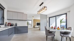 A modern kitchen with grey cabinets, a dining area with a white table and grey chairs, and a large window with a view of palm trees. - Landscape View by Homie (Ponta do Sol)
