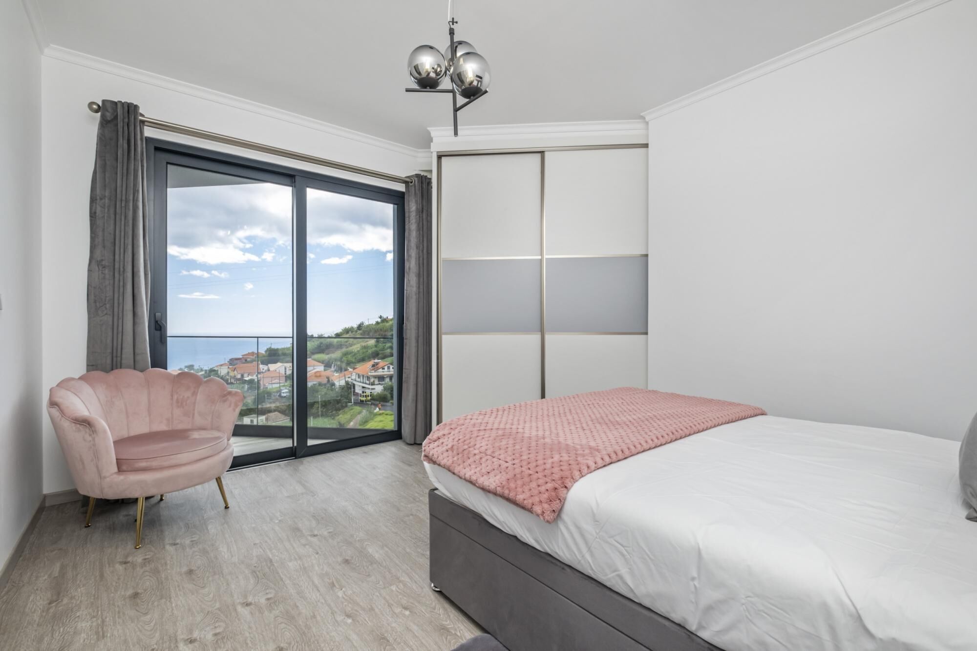 A bedroom with a bed, a chair, a wardrobe, and a view of a coastal town.