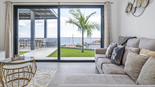 A modern living room with a large sliding glass door leading to a balcony with a view of the sea and a palm tree. - Landscape View by Homie (Ponta do Sol)