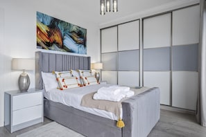 Un dormitorio moderno con una cama tapizada en gris, ropa de cama blanca y almohadas decorativas. Hay dos mesitas de noche con lámparas, un cuadro abstracto grande sobre la cama y un armario con puerta corredera de vidrio.