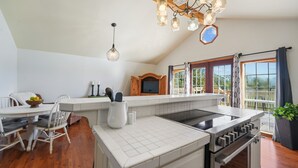 A modern kitchen with a central island, a dining area with a white table and chairs, and a large window with a view of the outdoors.