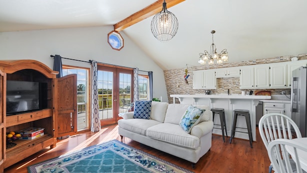 A spacious living room with a white sofa, a wooden entertainment unit, and a large TV. There is a kitchen with white cabinets and a stone backsplash. The room has a high ceiling with exposed beams and a chandelier.