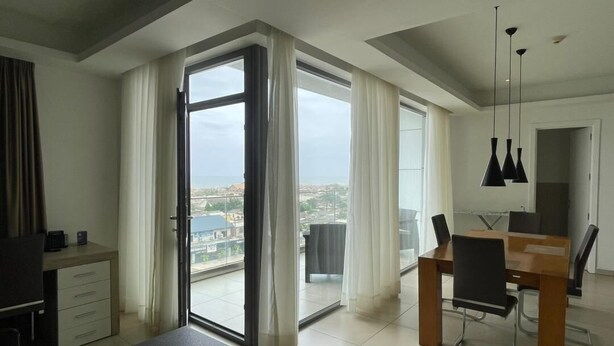 A modern hotel room with a balcony, a dining area, and a view of the city. - Apartment With Ocean View (Accra)