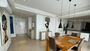 A modern dining area with a wooden table and chairs, a kitchen in the background, and artwork on the walls. - Apartment With Ocean View (Accra)