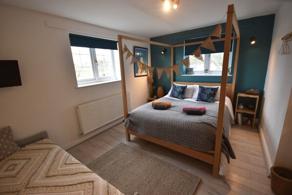 A bedroom with a four-poster bed, a window with blue curtains, a wooden floor, and a small bedside table with a lamp.
