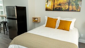 Una habitación de hotel con una cama, un refrigerador negro, una mesita de noche de madera y un cuadro de un bosque.