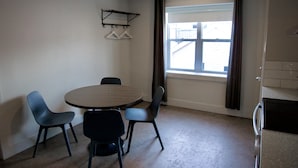 Una habitación con una mesa redonda, cuatro sillas, una ventana con cortinas y un estante con perchas en la pared.