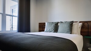 Un letto rifatto con cura, con una coperta a motivi scuri e due cuscini, una testiera in legno e una finestra con vista sugli edifici.