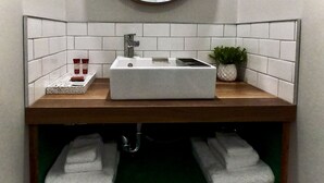 Un bagno con un lavabo rettangolare, uno specchio rotondo, piastrelle bianche, una piccola pianta in vaso e due asciugamani su uno scaffale.