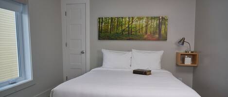 A bedroom with a large bed, a window with blinds, a door, a painting of a forest, and a bedside table with a lamp.