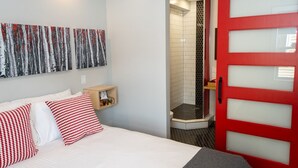 Habitación de hotel con una cama, almohadas estampadas en rojo y blanco, un estante de madera con objetos, una puerta roja y un área de ducha con azulejos blanco y negro.