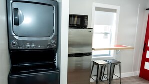 Cocina compacta con horno integrado, microondas y refrigerador de acero inoxidable. Hay un taburete pequeño y una ventana que proporciona luz natural.