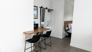 Una habitación compacta con un escritorio de madera, dos sillas negras, una lavadora blanca y una cama con ropa blanca.