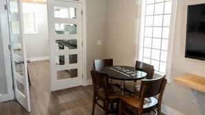 Un coin repas avec une table ronde et quatre chaises, une porte blanche avec des panneaux de verre et une fenêtre avec des stores.