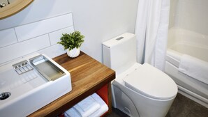 Un baño con inodoro, lavamanos con dispensador de jabón, una pequeña planta en maceta y bañera con cortina de ducha.