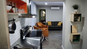 Una cocina moderna con estufa, lavamanos y una silla naranja. Se ve un sofá al fondo y un pequeño estante con una planta.