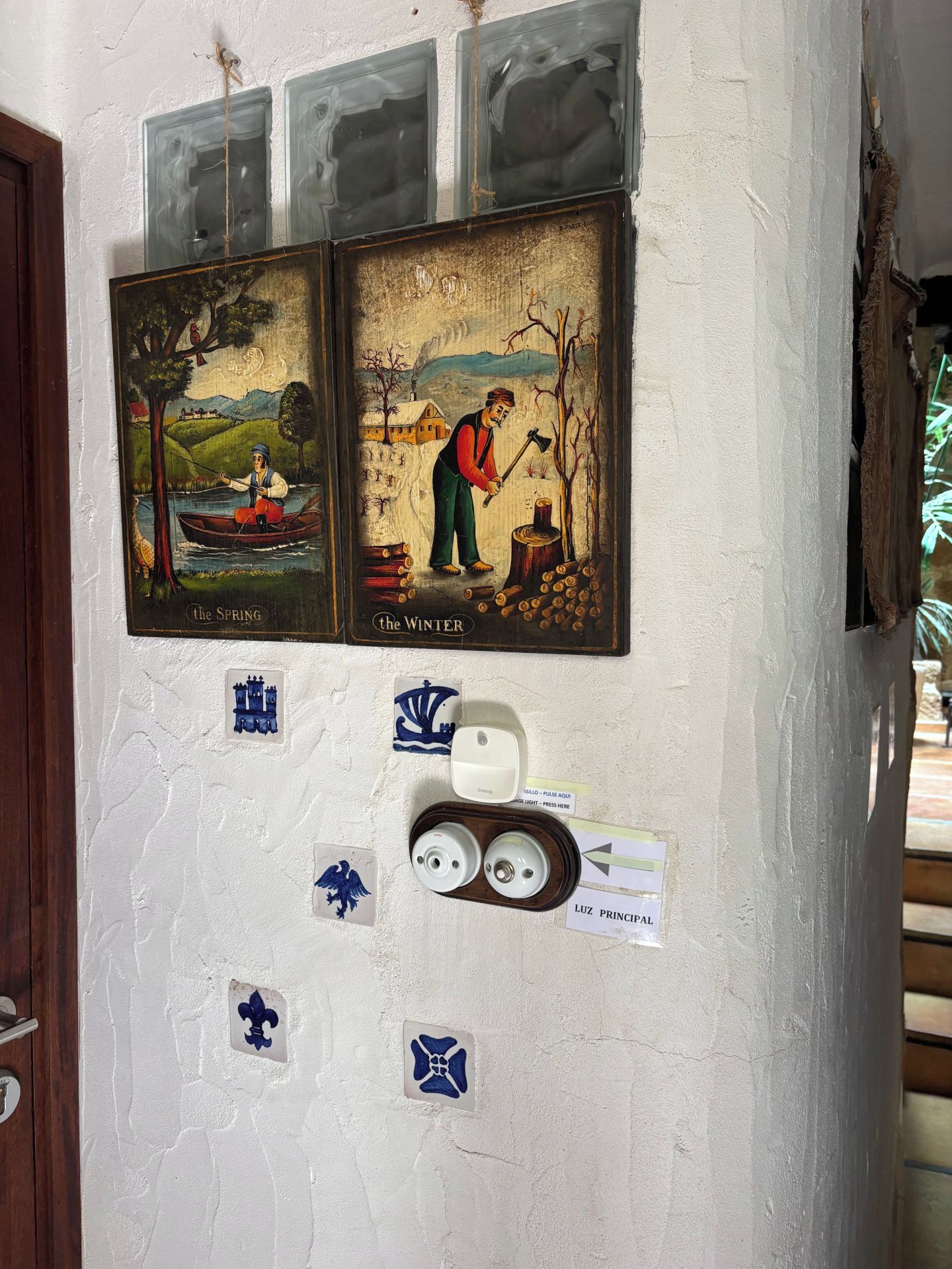 A wall with three framed paintings depicting scenes labeled 'the SPRING' and 'the WINTER,' a wall-mounted light switch, and several small blue and white tiles.
