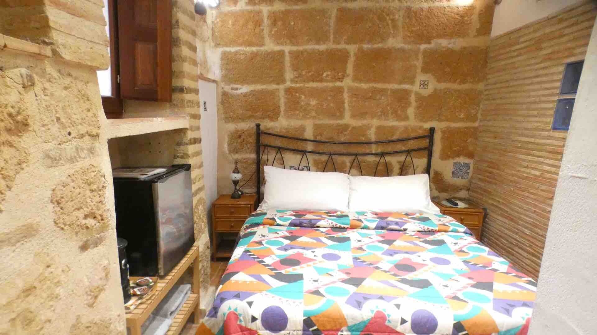 A small, cozy room with a bed, a nightstand, a wooden shelf, and a stone wall.