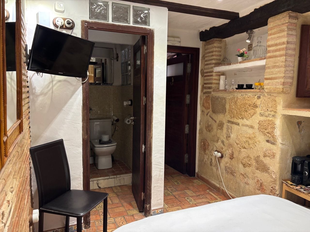 Una habitación con cama, una silla, un televisor y un baño con inodoro, además de una pared de piedra.