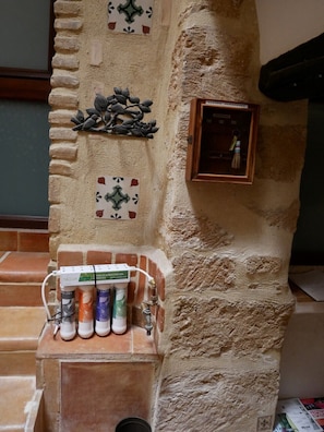 Eine Steinsäule mit einem verzierten Metallteil und einer kleinen Holzschatulle. Auf einer gefliesten Ablage stehen drei Wasserfilter.