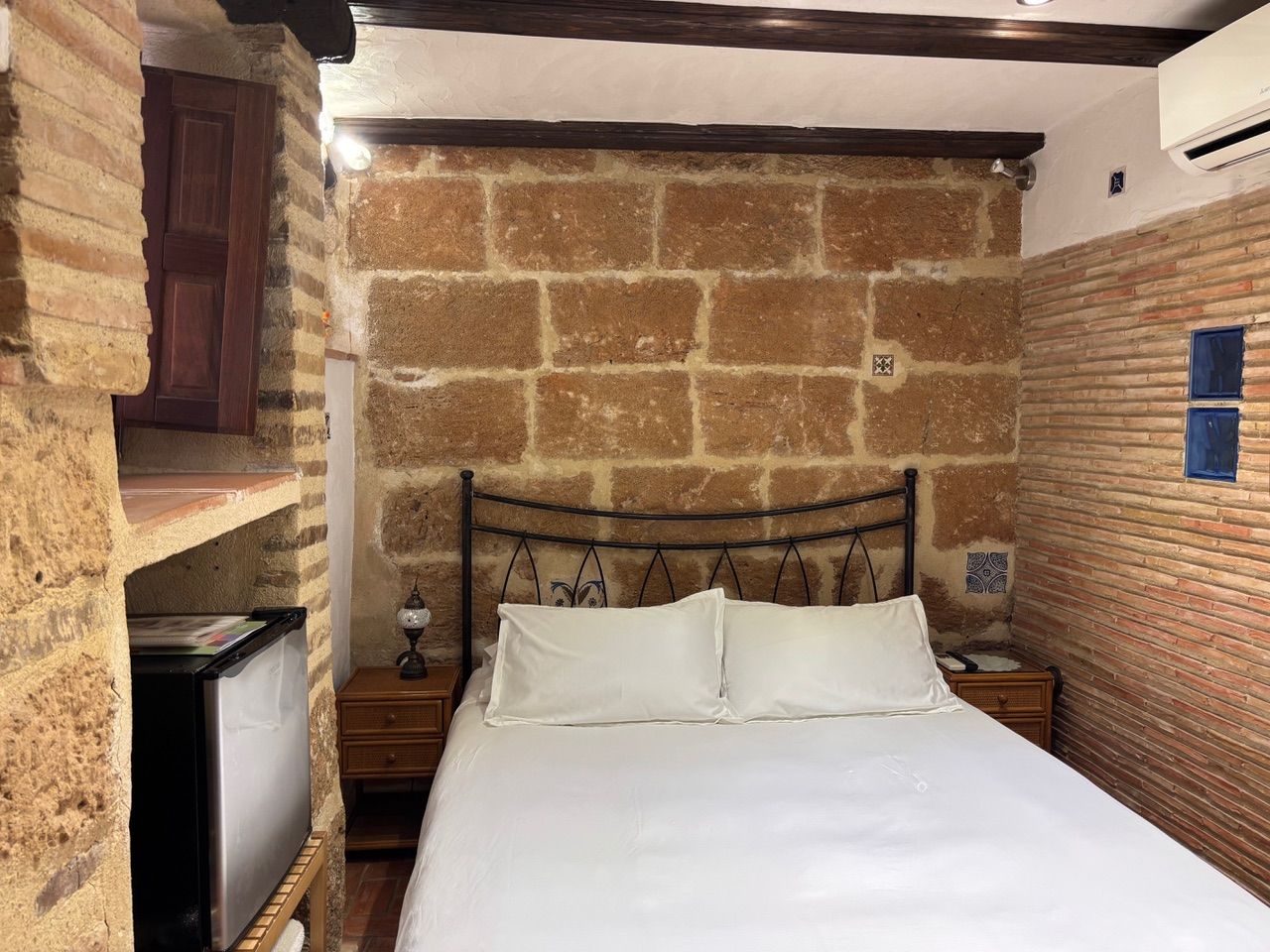 Una cama individual con ropa de cama blanca en una habitación con paredes de ladrillo visto y techo de madera.