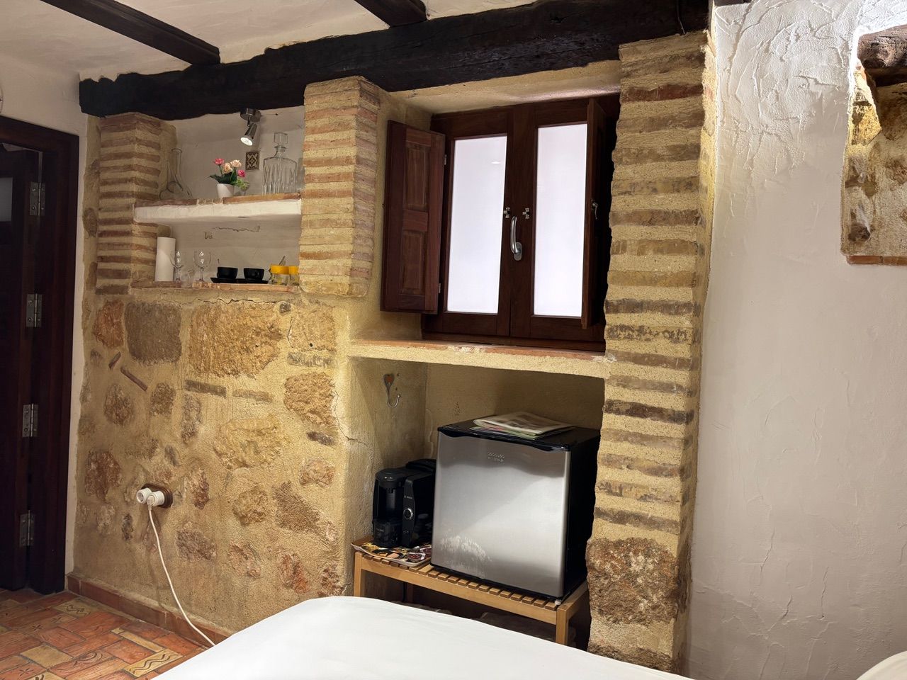 A room with a stone wall, a wooden shelf with a coffee maker, and a white bed.