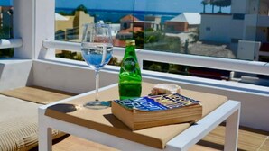 Een tafel met een glas blauwe vloeistof, een groene fles en een boek op een balkon met uitzicht op gebouwen en de zee.