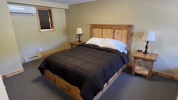 A bedroom with a wooden bed, two bedside tables with lamps, a window with blinds, and an air conditioning unit.