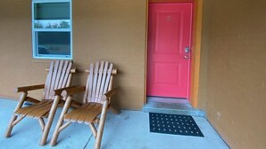 Un bâtiment avec une porte rouge, deux chaises en bois à l’extérieur et une petite fenêtre.