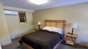 Ein Schlafzimmer mit einem hölzernen Bett, zwei Nachttischen mit Lampen, einem Fenster mit Jalousien und einer Klimaanlage.