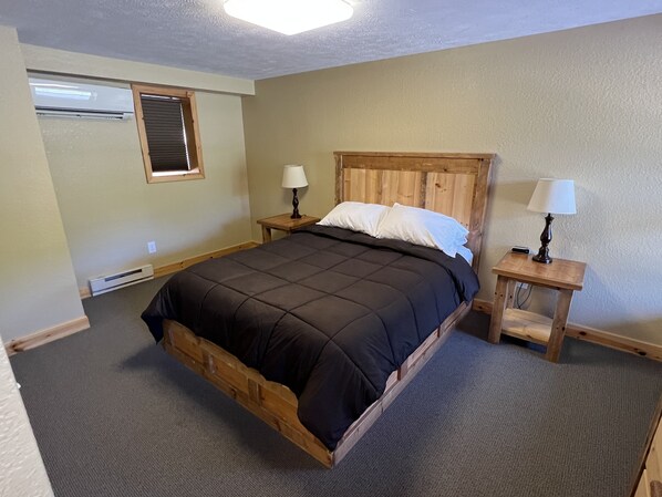 Une chambre à coucher avec un lit en bois, deux tables de chevet avec des lampes, une fenêtre avec des stores et un appareil de climatisation.