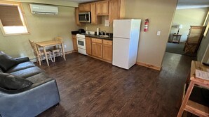 Una cocina compacta con armarios de madera, un refrigerador blanco y un microondas. Hay una mesa de comedor con sillas y un sofá gris.