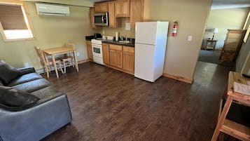 A compact kitchen with wooden cabinets, a white refrigerator, and a microwave. There is a dining table with chairs and a gray sofa.