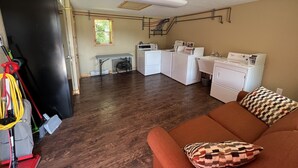 Ein Waschraum mit Waschmaschine, Trockner und Bügelecke. Es gibt einen Holzboden und ein braunes Sofa mit einem gemusterten Kissen.