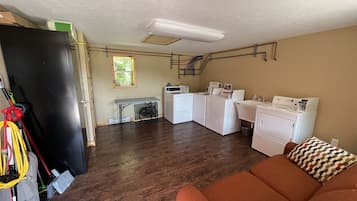A laundry room with a washing machine, dryer, and ironing station. There is a wooden floor and a brown couch with a patterned pillow.