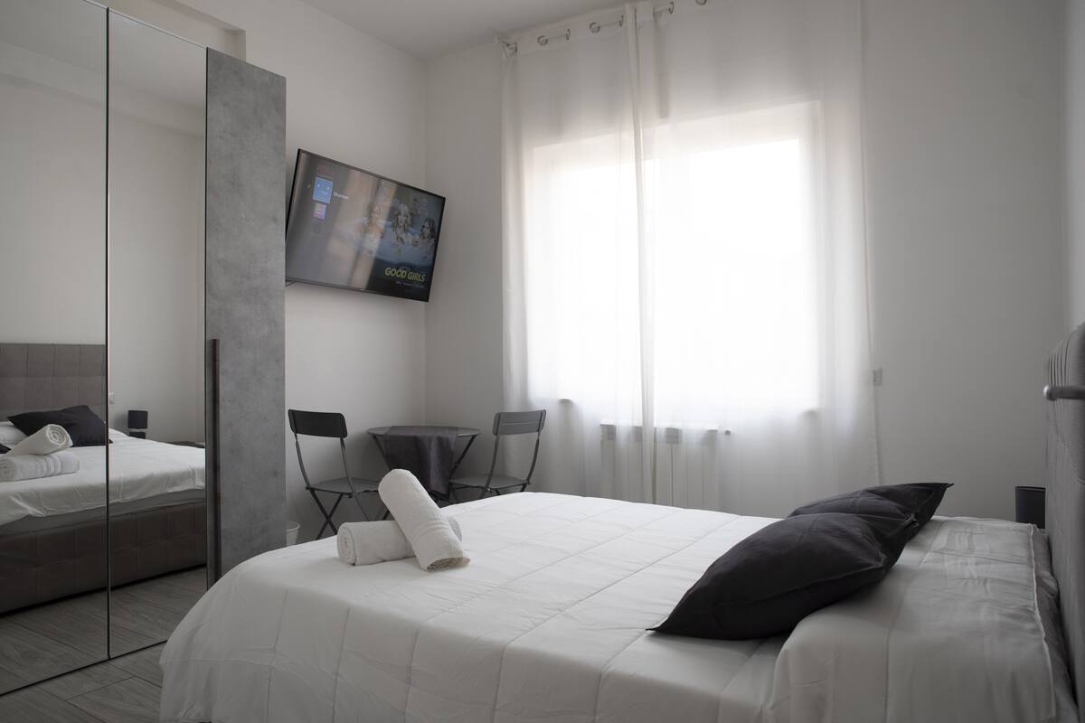 A modern hotel room with a large bed, a flat-screen TV mounted on the wall, a seating area with a table and chairs, and a large window with sheer curtains.