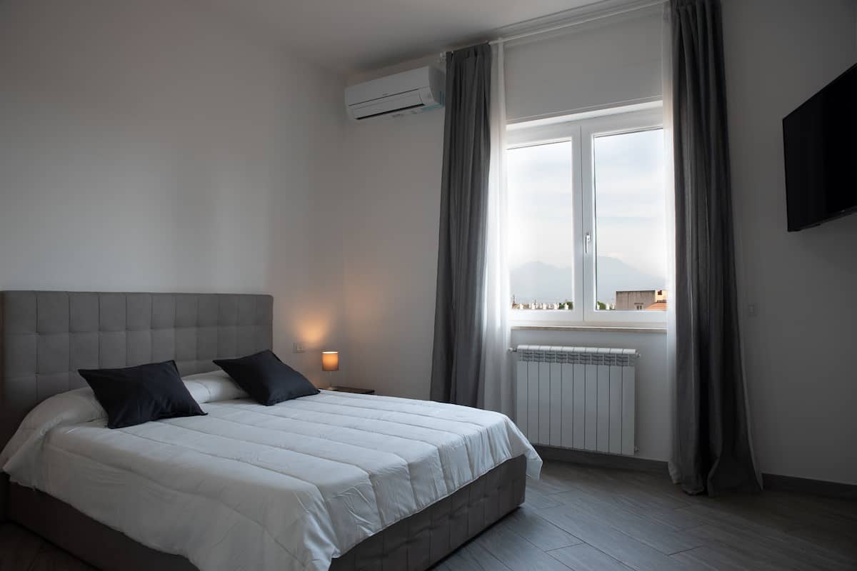 A bedroom with a large bed, a nightstand, an air conditioning unit, a window with curtains, and a wall-mounted TV.