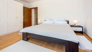 A modern bedroom with a large bed, wooden flooring, white walls, and a wooden wardrobe.