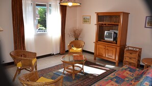 Ein Wohnzimmer mit Rattanmöbeln, einem Glastisch, einem Fernseher auf einer Holzkommode und Blick durch ein mit Vorhängen drapiertes Fenster.