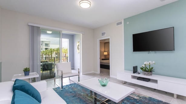 Een moderne woonkamer met een witte bank, kussens in de kleur turquoise, een glazen salontafel en een flatscreen televisie die aan de muur is bevestigd.