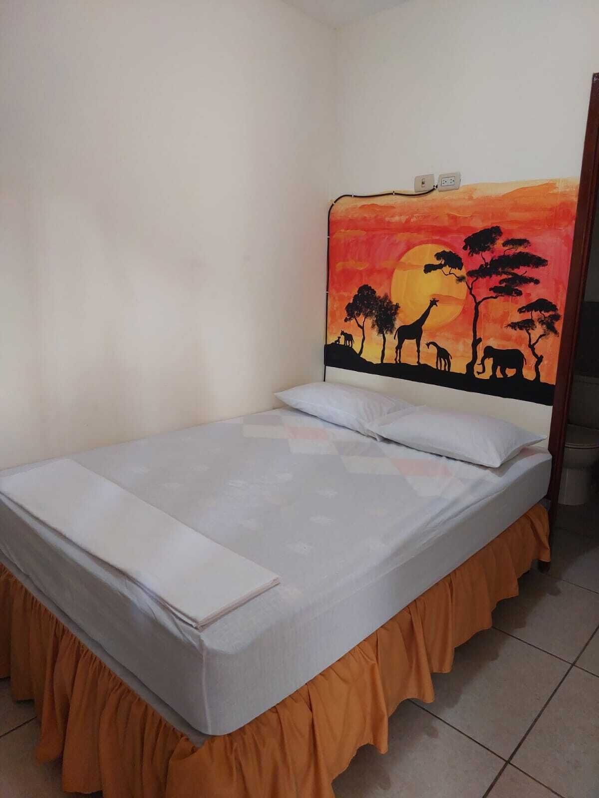 A single bed with a white mattress and orange skirted legs, a plain wall, and a sunset painting above the bed.