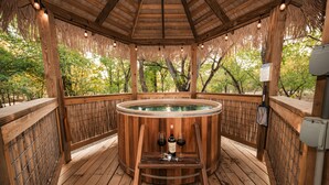 A wooden hot tub with two bottles of wine and a wine glass on a wooden platform.