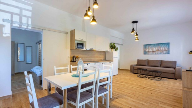 A modern kitchen with white cabinets, a microwave, and a sink. There is a dining area with a white table and chairs. A brown sofa is against the wall, and there are two small tables with chairs.