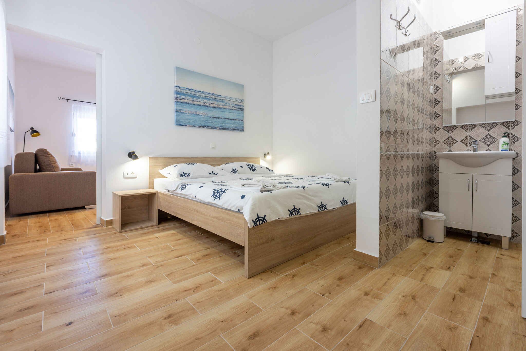 Um quarto com cama de madeira, mesa de cabeceira, abajur, prateleira fixada na parede, janela com cortinas e banheiro com pia e espelho.
