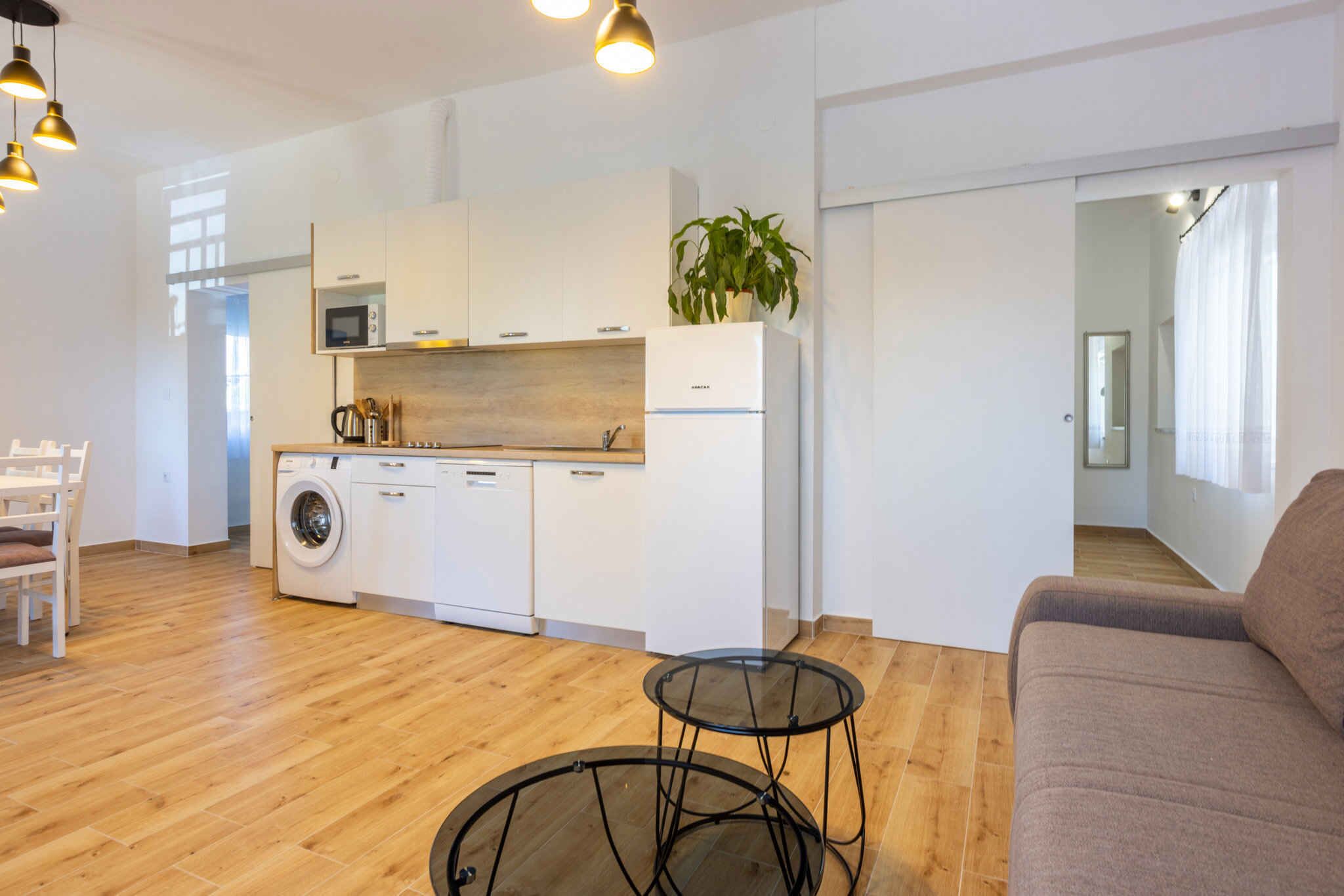 A modern kitchen with white cabinets, a microwave, a washing machine, and a plant. There are two glass tables and a sofa in the living area.