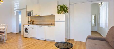 A modern kitchen with white cabinets, a microwave, a washing machine, and a plant. There are two glass tables and a sofa in the living area.