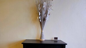 A small, dark wooden table with a vase of dried flowers on top, flanked by two metal chairs with patterned cushions, against a plain wall.