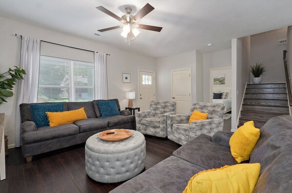 A living room with a grey sofa, a grey ottoman, a grey armchair, and a wooden coffee table. There is a ceiling fan, a window with blinds, and a staircase in the background.