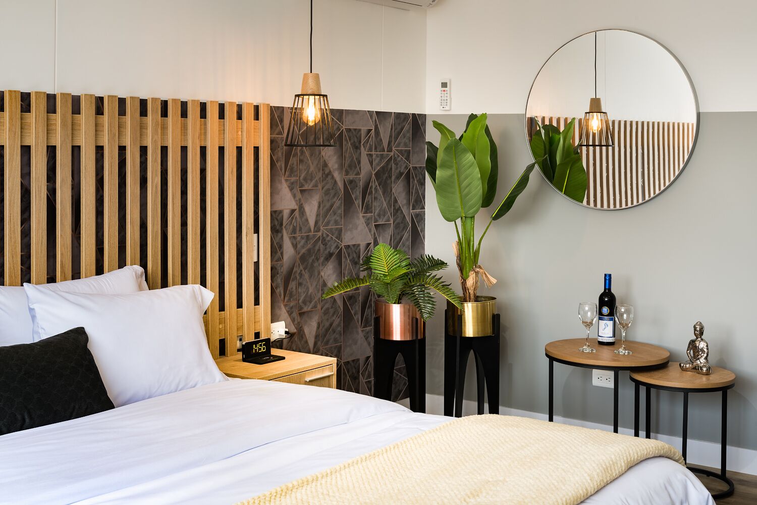 A modern hotel room with a wooden headboard, a large bed, a nightstand with a clock, a round mirror, a small table with a wine bottle and glasses, and two potted plants.