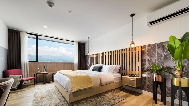A modern hotel room with a large bed, a desk, a chair, a potted plant, and a view of the mountains. - Go Living and Suites by Jalo (Medellín)